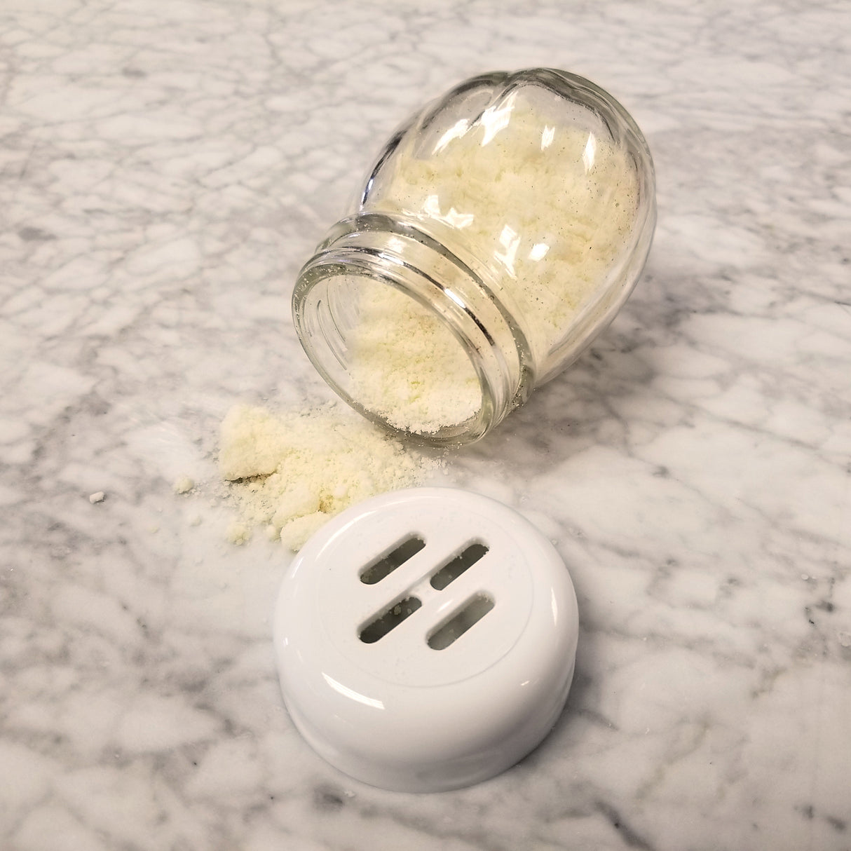 A swirl glass shaker jar, likely a 6 oz replacement jar, is tipped over on a gray marble surface, spilling white powder. The shaker’s white plastic lid with three long holes is detached and lies in the foreground.