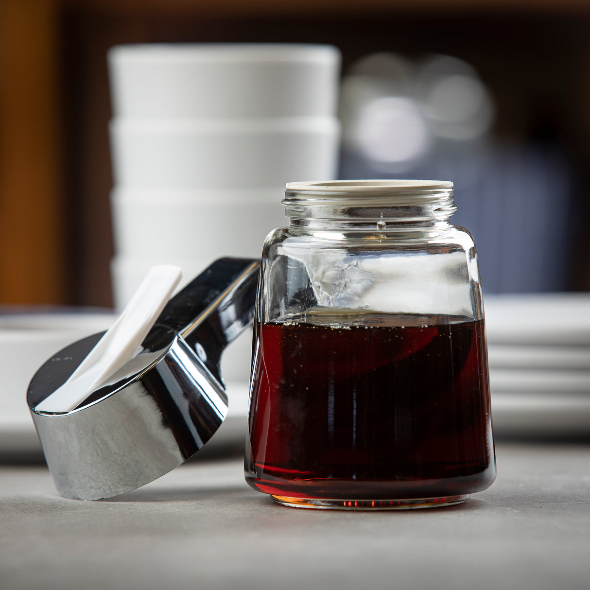 A Modern Glass Dispenser filled with dark syrup sits on a gray surface. The metal lid, chrome plated and open, rests nearby. Behind the jar, stacked white ceramic cups and blurred plates create a clean, café-like setting.