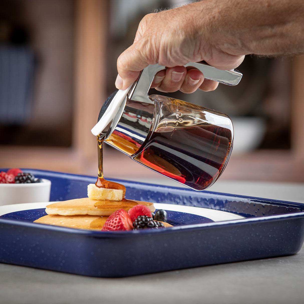 A hand pours syrup from a Modern Glass Dispenser with a chrome plated top onto pancakes stacked on a blue-rimmed plate, joined by fresh strawberries, blackberries, and blueberries. The scene sits on a gray countertop with a bowl of more berries in the background.