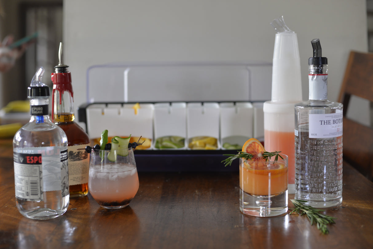 A wooden table with bottles of liquor featuring BPA free plastic cups, two colorful cocktails garnished with herbs and fruit, a container of sliced citrus, a jigger, a bitters bottle, and bottles equipped with sanitary spouts. The background is softly blurred.