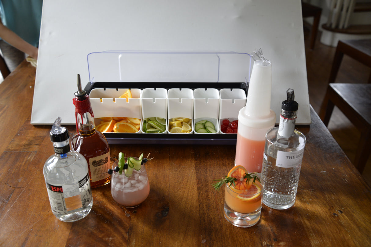 A wooden table displays cocktail ingredients: bottles of alcohol, two mixed drinks, a juice bottle with a sanitary spout, cut fruits and garnishes, plus bar tools. Sliced citrus, cucumbers, and cherries are visible; drinks are garnished with rosemary and lime.