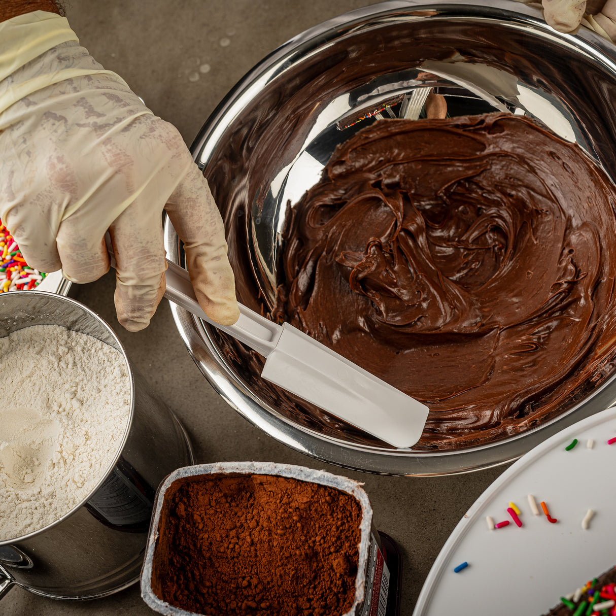 A gloved hand holds a BPA free white spatula, mixing glossy chocolate batter in a metal bowl. Nearby are a container of cocoa powder, a bowl of flour, and colorful sprinkles scattered on a gray surface.