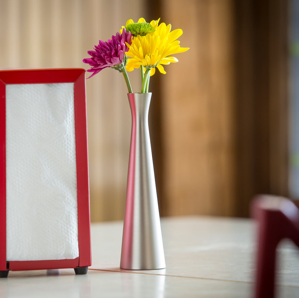 A zinc alloy Flower Vase with a brushed finish holds yellow, purple, and green flowers on a table beside a red napkin holder filled with white napkins. The softly blurred background highlights warm, wooden tones.