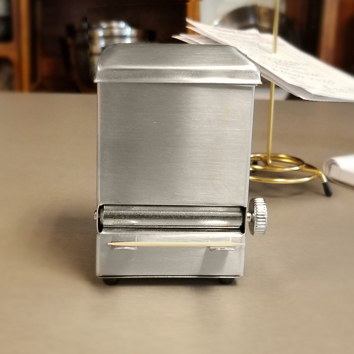 A small, rectangular stainless steel toothpick dispenser with a top lid, a front roller featuring a ridged knob on the right, and a toothpick in front. The blurry kitchen counter background includes a gold order holder and scattered papers.