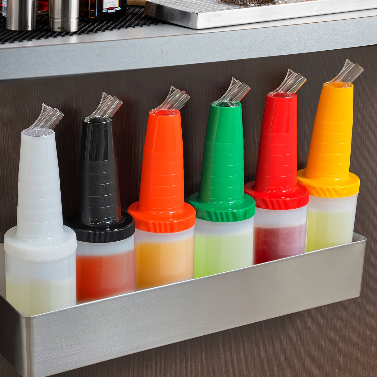 Six Quart Pourmaster condiment bottles with assorted color storage caps and polyethylene pour spouts are lined up in a metal rack under a counter, perfect for organizing syrups or sauces in any bar or kitchen setting.