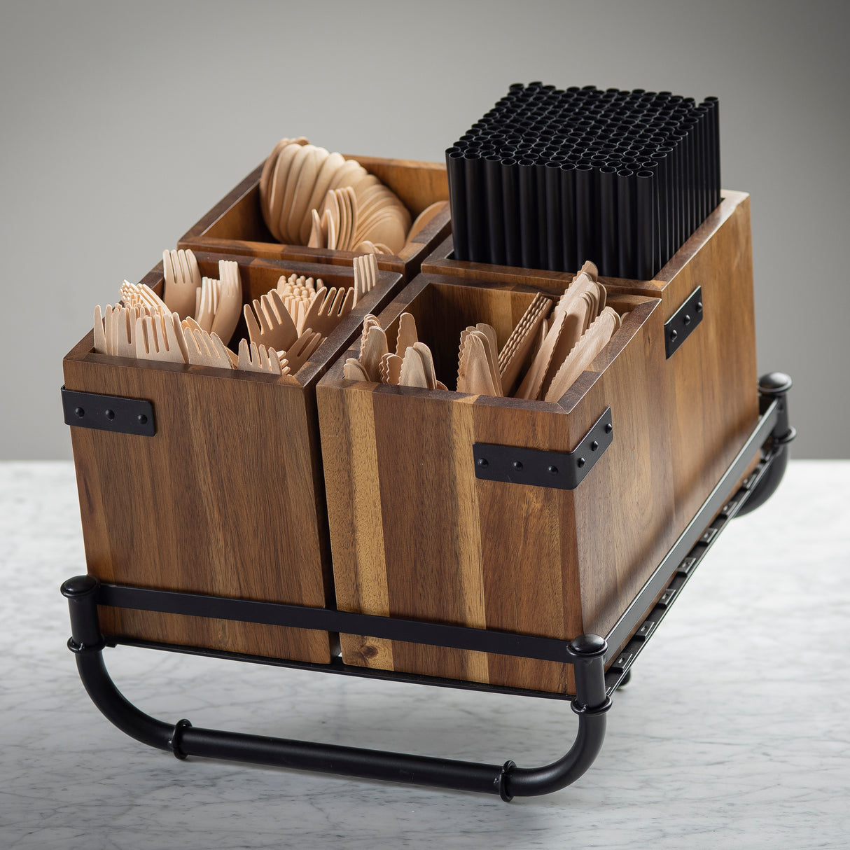 A wooden cutlery holder rack with four compartments holds wooden spoons, forks, and knives, plus a section for stacked black plastic straws. The set features black metal accents and base, blending seamlessly into any industrial collection.