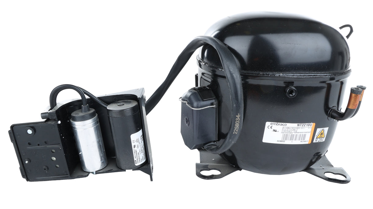 An Empura Refrigeration compressor, model E-EGM-23FB, with attached capacitors and wiring sits on a white background. The cylindrical unit features metal mounting brackets, a visible label, and a separate black bracketed capacitor housing.