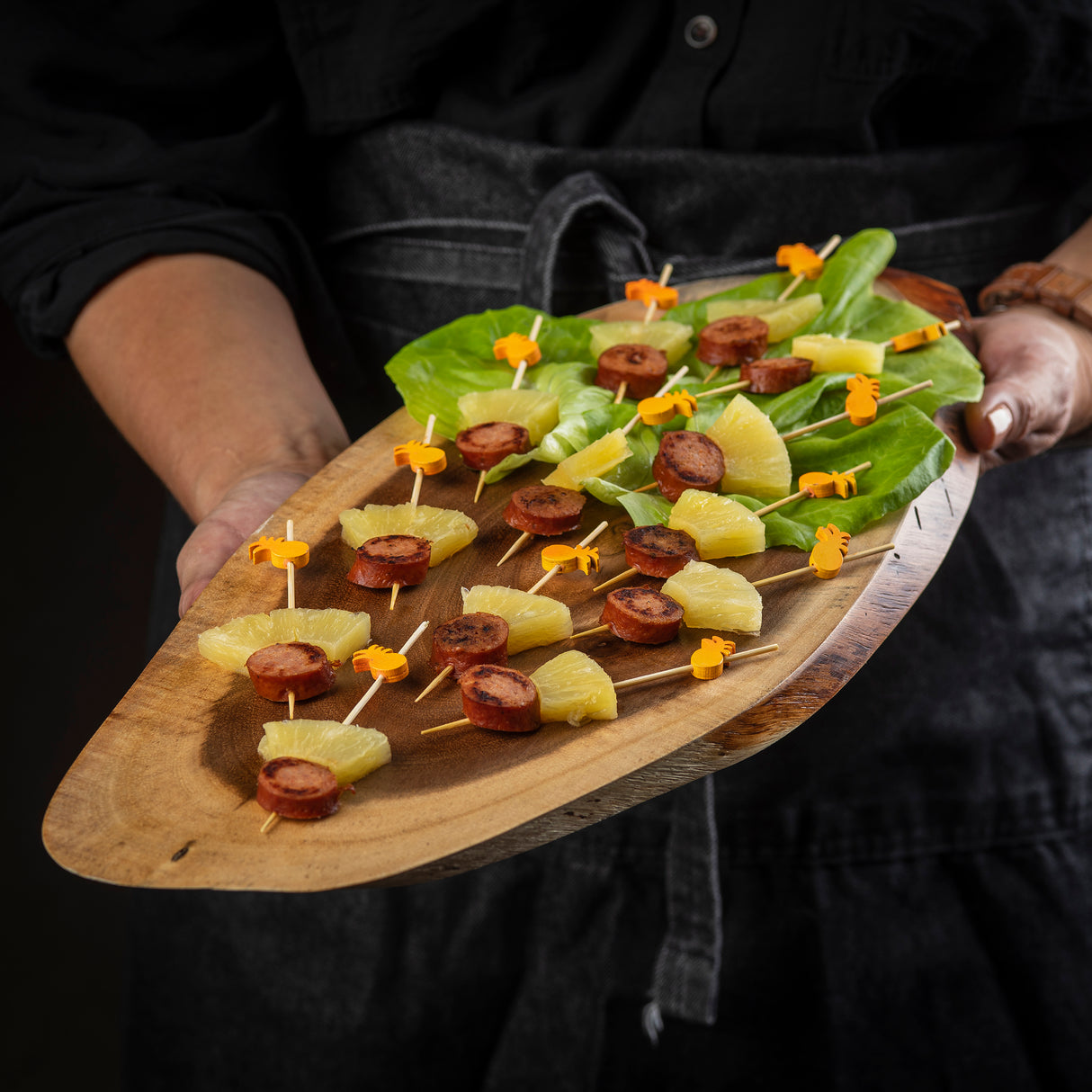 A person in dark clothing and a gray apron holds a TableCraft Products Acacia Serving Board topped with skewers of sausage and pineapple, each secured with an orange animal-shaped pick and resting on fresh green lettuce leaves.