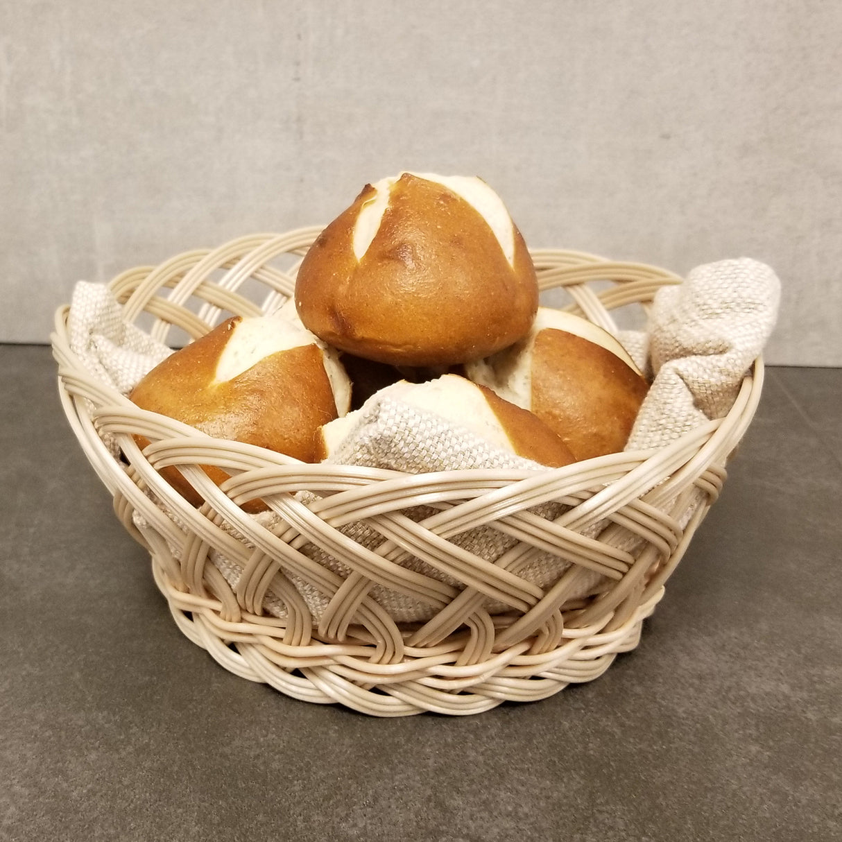 A hand-woven polypropylene basket filled with four brown, round bread rolls with white tops. A light tan textured napkin lines the basket, which sits on a dark gray surface against a plain beige background.