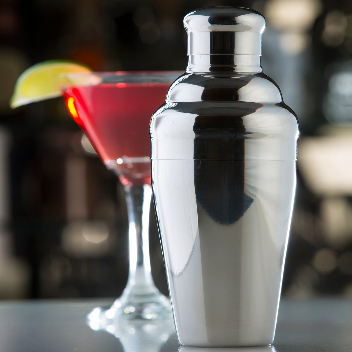 A shiny stainless steel cocktail shaker from a premium bar shaker set stands in the foreground, reflecting light. In the blurred background, a martini glass filled with a red cocktail and garnished with lime suggests a lively cocktail lounge.
