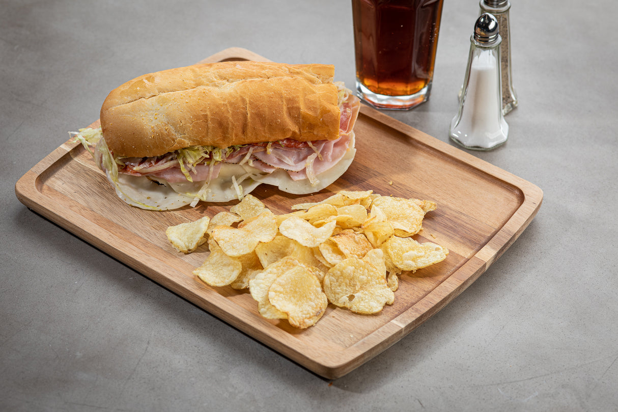 A wood serving plate holds a sub sandwich with deli meats, cheese, shredded lettuce, and a side of potato chips. Behind the tray is a glass of iced tea and salt and pepper shakers on a plain, light-colored tabletop.
