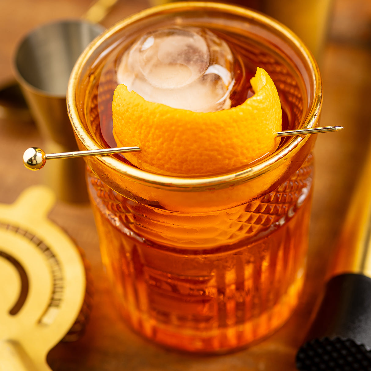 A close-up of an amber cocktail in a textured glass, garnished with an orange peel held by a gold cocktail pick. Stainless steel and gold bar tools surround the glass on a wooden surface, enhancing the elegant presentation.