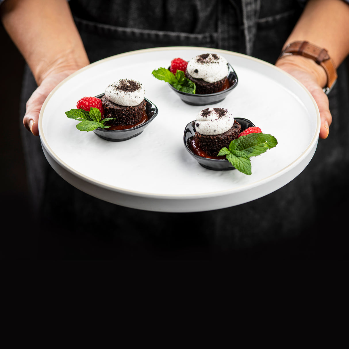 A person holds a white melamine serving tray with three small chocolate desserts topped with whipped cream, cookie crumbs, a mint sprig, and a raspberry in black bowls. Both the tray and bowls are dishwasher safe. The background and outfit are dark.
