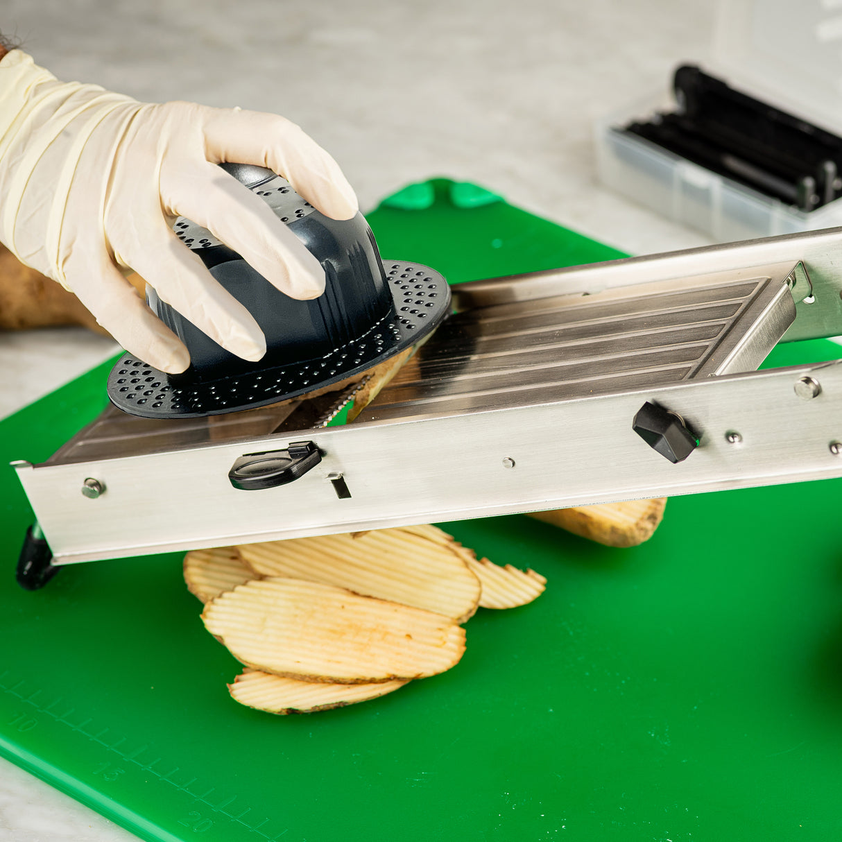 A gloved hand uses a TableCraft stainless steel mandoline slicer to slice a potato on a green cutting board. Thin, crinkle-cut potato slices are collected below the slicer, with various kitchen tools visible in the background.
