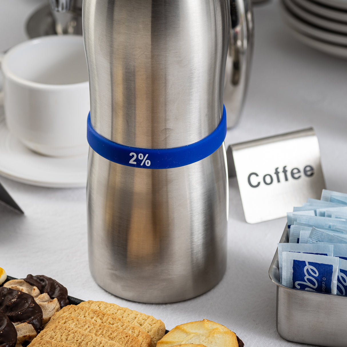 A stainless steel carafe labeled 2% with a blue Silicone Flavor Band stands on a white tablecloth beside a sign reading Coffee, TableCraft Products plates, a cup and saucer, packets of sugar, and an assortment of cookies and pastries.