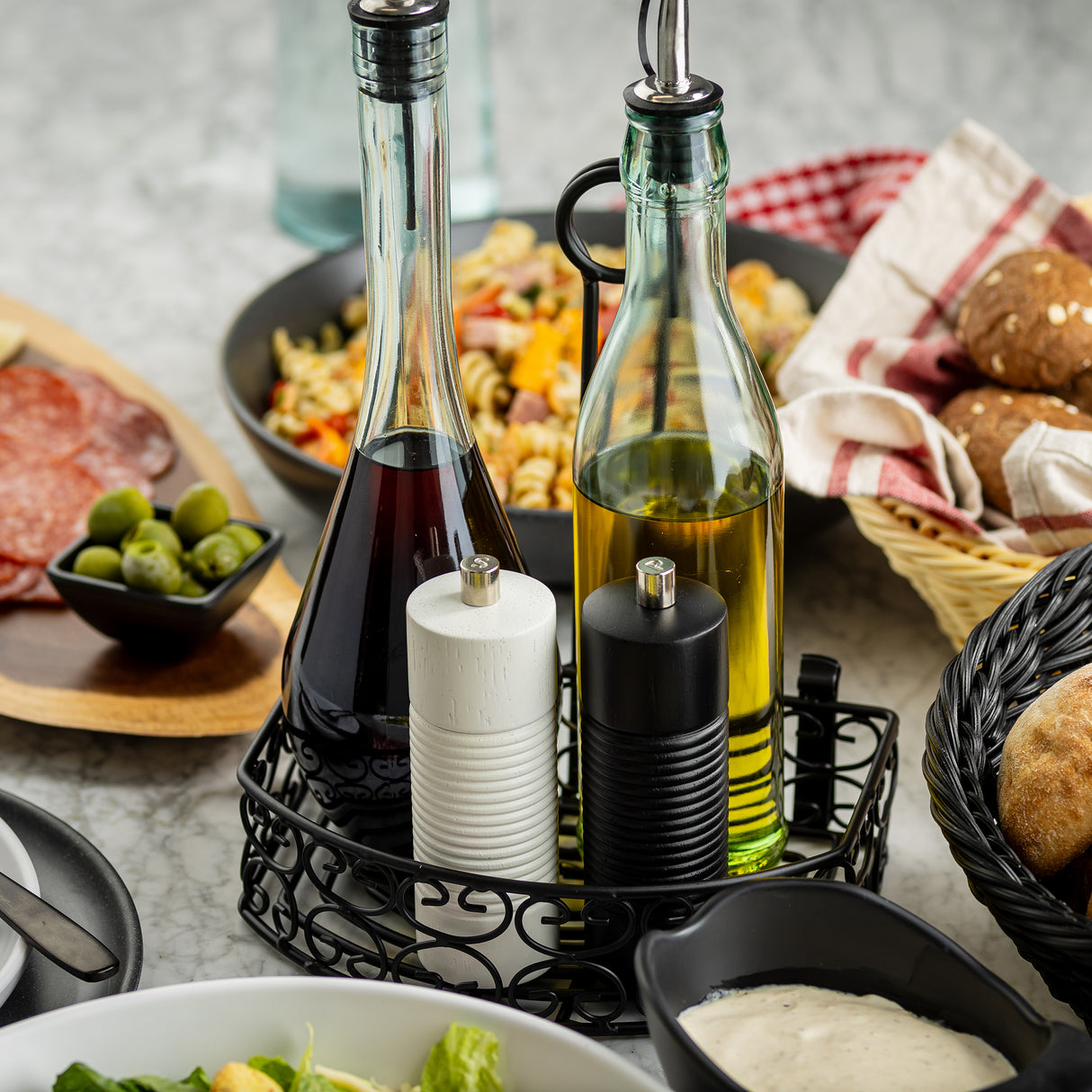 Two glass bottles of vinegar and oil, with a stylish wood salt and pepper grinder from TableCraft Products, sit in a black metal holder. Surrounding dishes include salad, bread rolls, green olives, charcuterie, and pasta on a marble table.