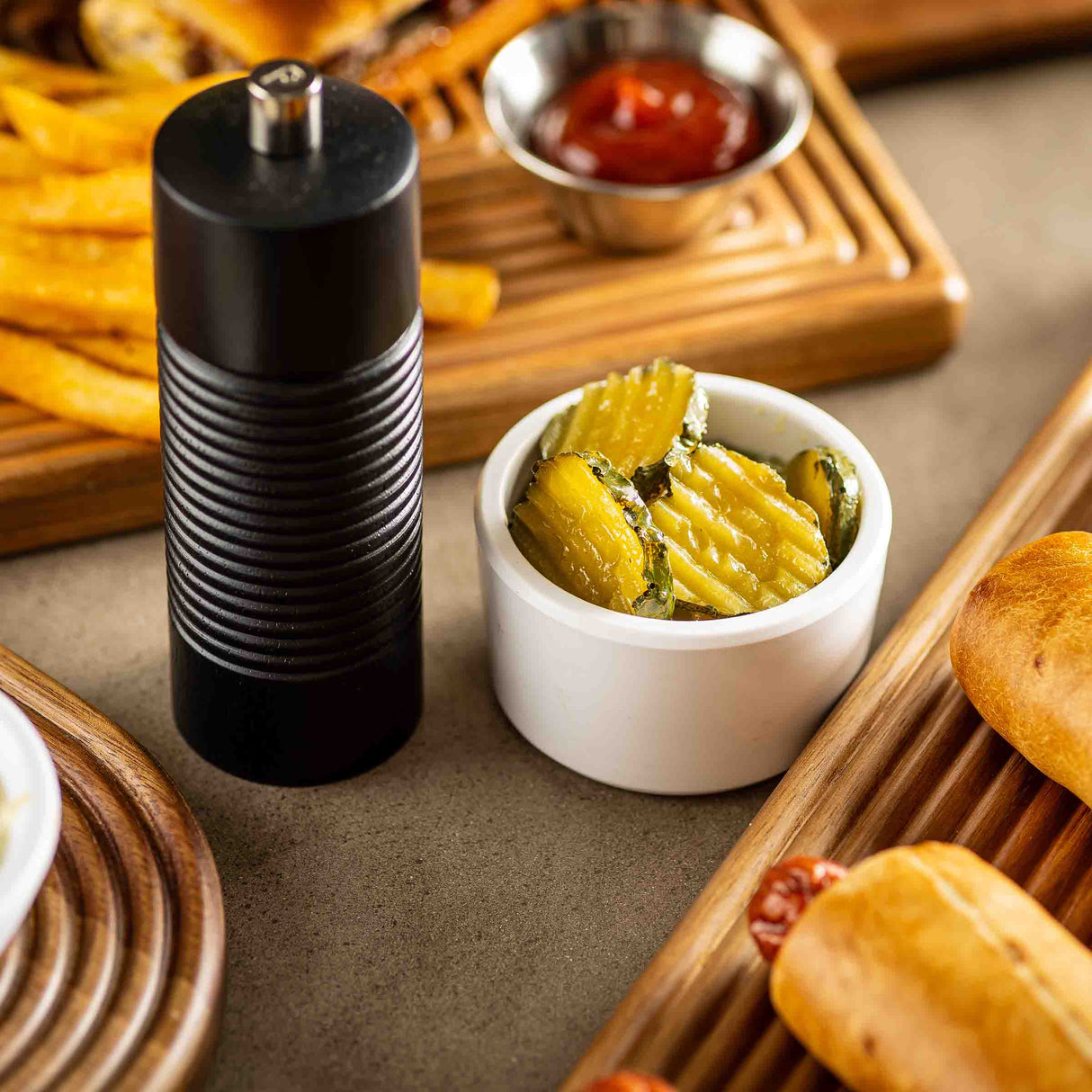 A TableCraft Products wood salt and pepper grinder sits next to a small white bowl of sliced pickles. In the background, crinkle-cut fries, ketchup, and part of a baked sausage roll on a wooden tray are visible.