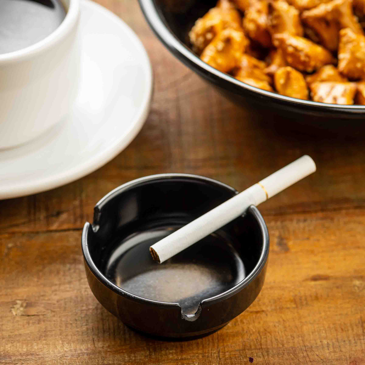 A close-up of a black phenolic ashtray holding a single unlit cigarette on a wooden table. To the left, a white coffee cup and saucer sit, with a bowl of snacks—possibly pretzels—partially visible in the background.
