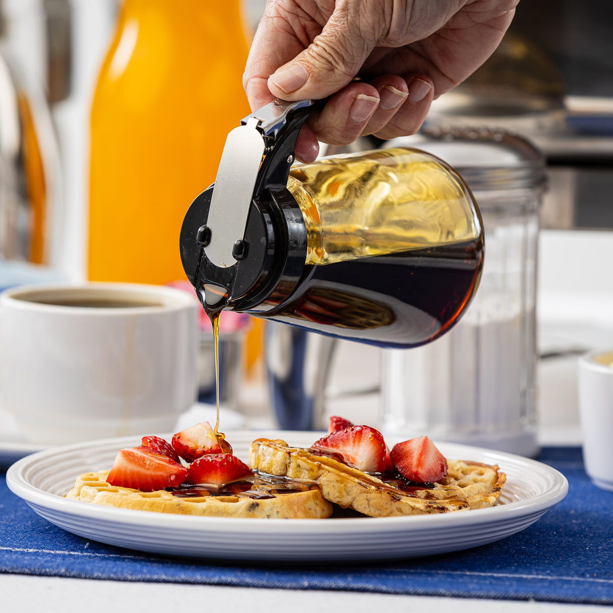 A hand pours syrup from a TableCraft Modern Glass Dispenser onto waffles topped with sliced strawberries on a white plate. A cup of coffee, a sugar container, and an orange bottle are in the background on a blue placemat.
