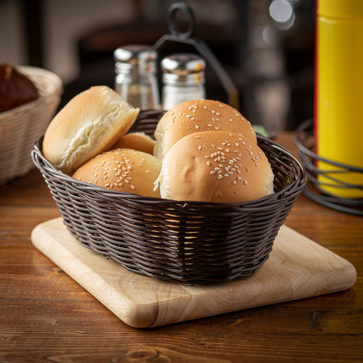 A hand-woven dark wicker basket holds four sesame seed hamburger buns, placed on a wooden cutting board. In the background are a metal condiment holder, salt and pepper shakers, and a yellow ketchup bottle on a cozy wooden table.
