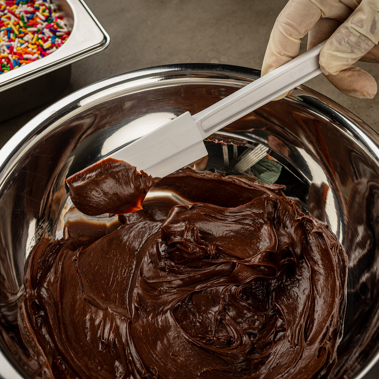 A gloved hand holds a BPA free spatula covered in rich, glossy chocolate frosting over a metal bowl filled with more frosting. In the background, a metal tray with colorful sprinkles sits on a gray countertop.