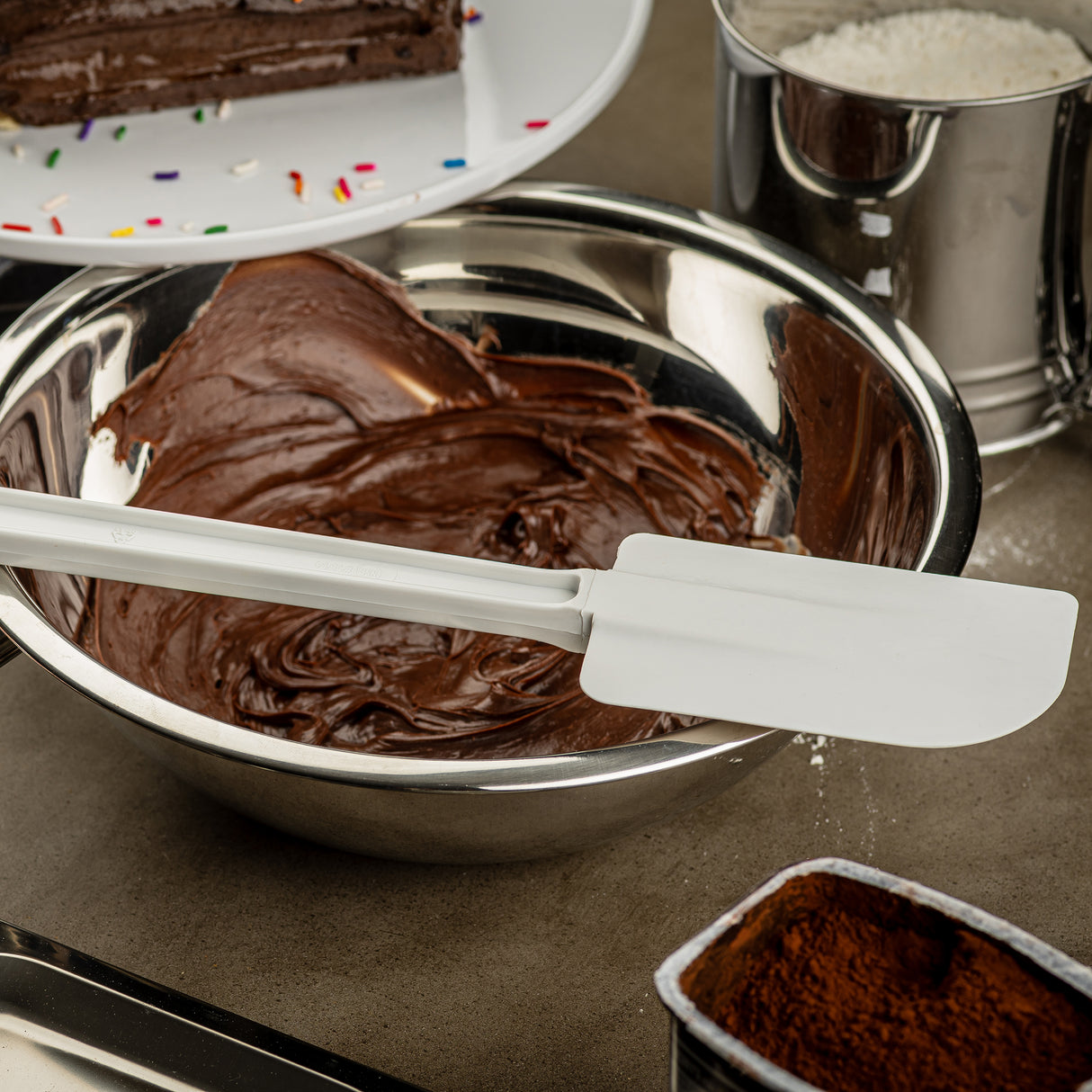 A metal bowl filled with chocolate frosting sits on a counter, with a BPA free, heat resistant spatula resting on its edge. Nearby are a partial chocolate cake on a plate, a metal sifter of flour, and an open container of cocoa powder.