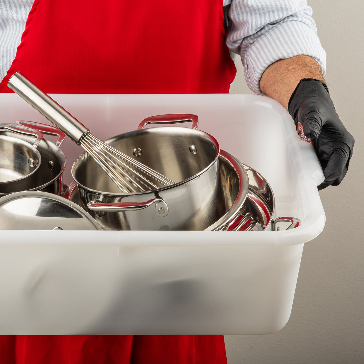 A person in a red apron and black glove holds a white tote box filled with clean, dishwasher safe stainless steel pots, pans, and a metal whisk. Only the torso and arm are visible against a plain, neutral background.