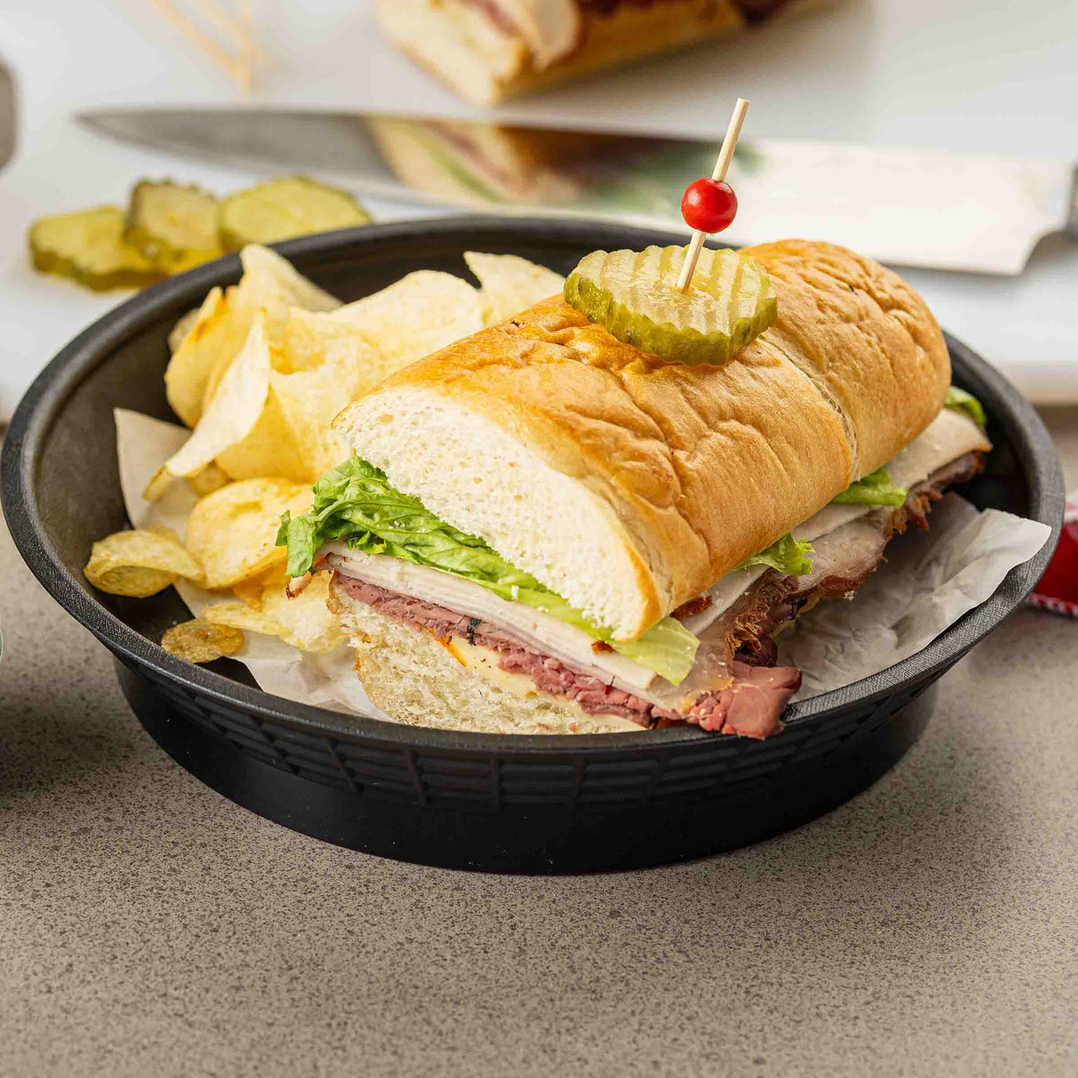 A sub sandwich with lettuce, meat, and cheese is served in a heat resistant black bowl with potato chips. The sandwich is topped with pickle slices held by a toothpick with a red bead. Pickle slices and a knife are visible in the blurred background.