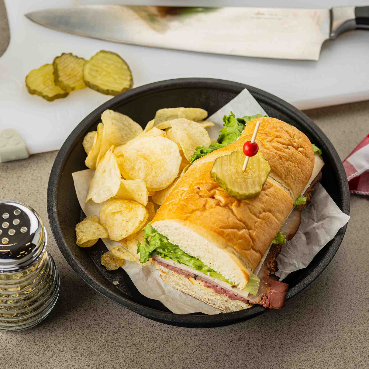A sandwich with lettuce, cheese, and deli meat on a hoagie roll, topped with a pickle slice and toothpick, sits in a dishwasher safe platter basket lined with parchment paper next to potato chips. A pepper shaker, pickle slices, and knife are in the background.