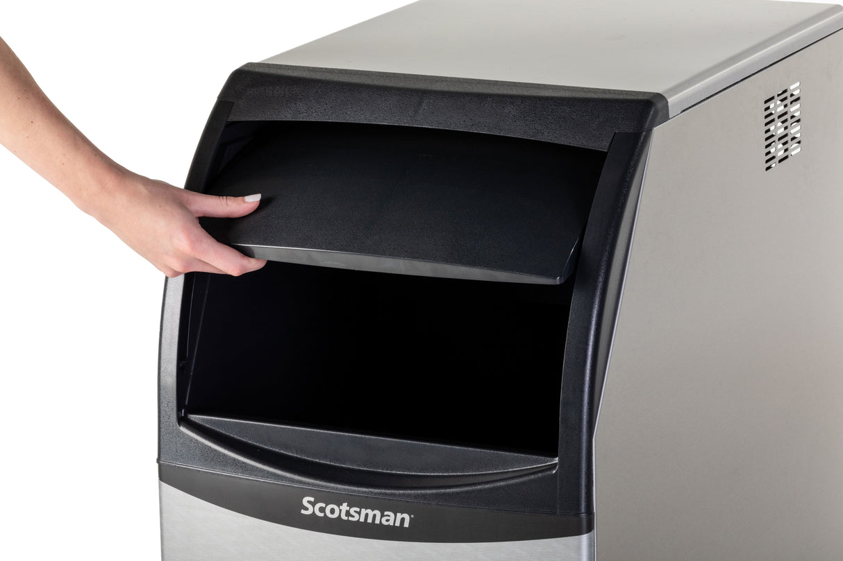A person’s left hand lifts the black lid of a stainless steel Scotsman Ice Maker, revealing the dark interior. This sleek, rectangular undercounter ice maker displays the Scotsman brand name prominently on the front.
