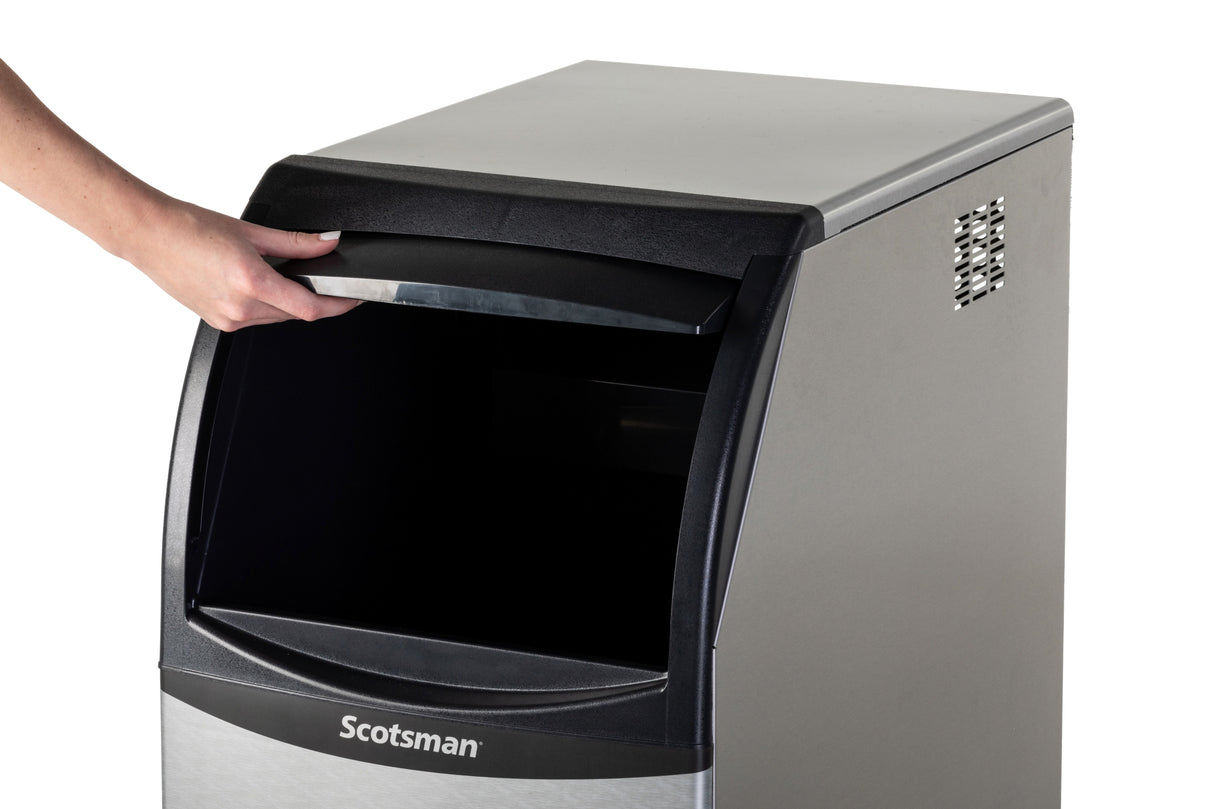 A person’s left hand lifts the black lid of a Scotsman undercounter ice maker. The machine has a silver and black exterior with ventilation holes on the right, and “Scotsman” on the front. The interior appears empty and unlit.