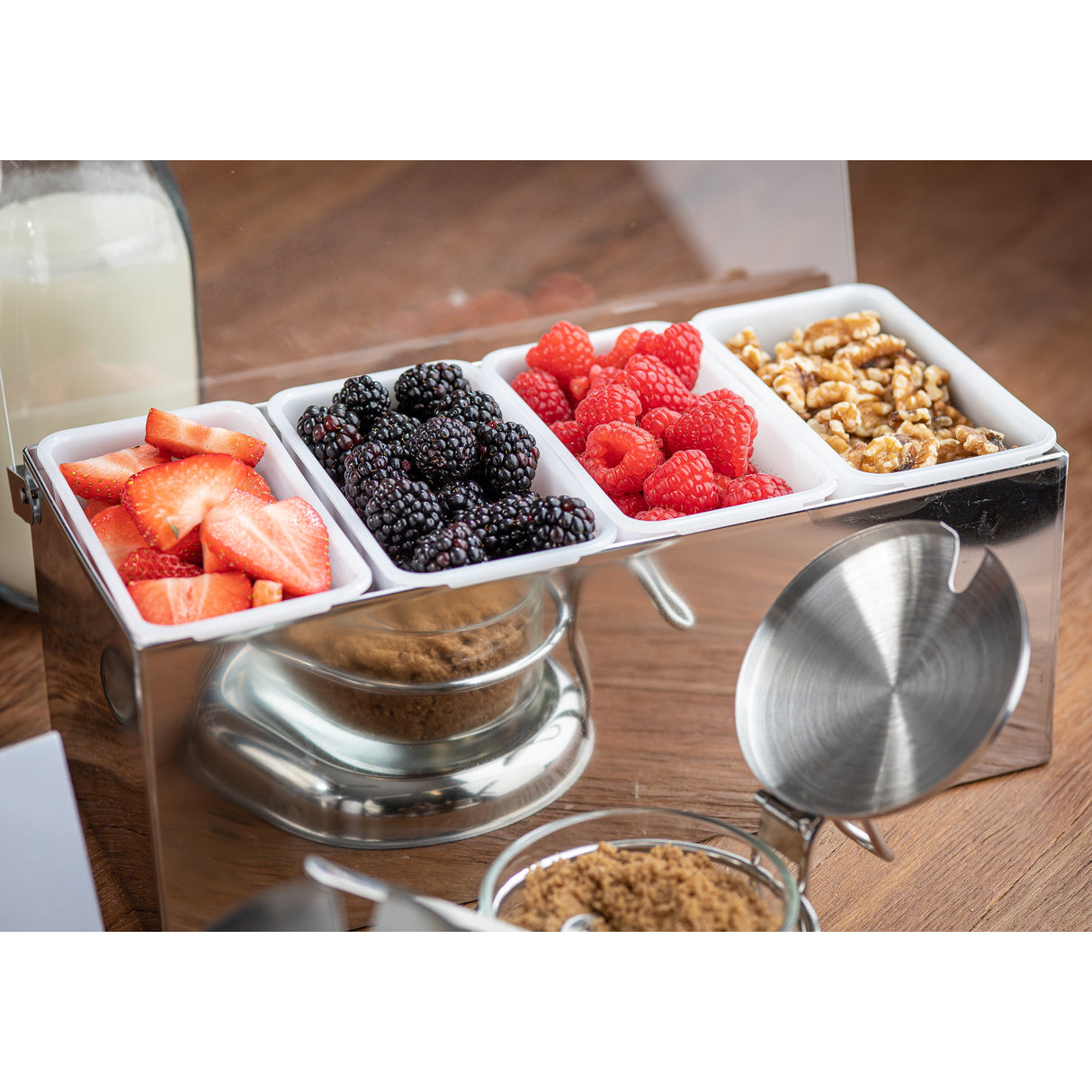 A close-up of four small white trays in a stainless steel condiment holder, filled with sliced strawberries, blackberries, raspberries, and walnut halves. A glass bottle of milk and jars of brown sugar sit nearby on a wooden table.