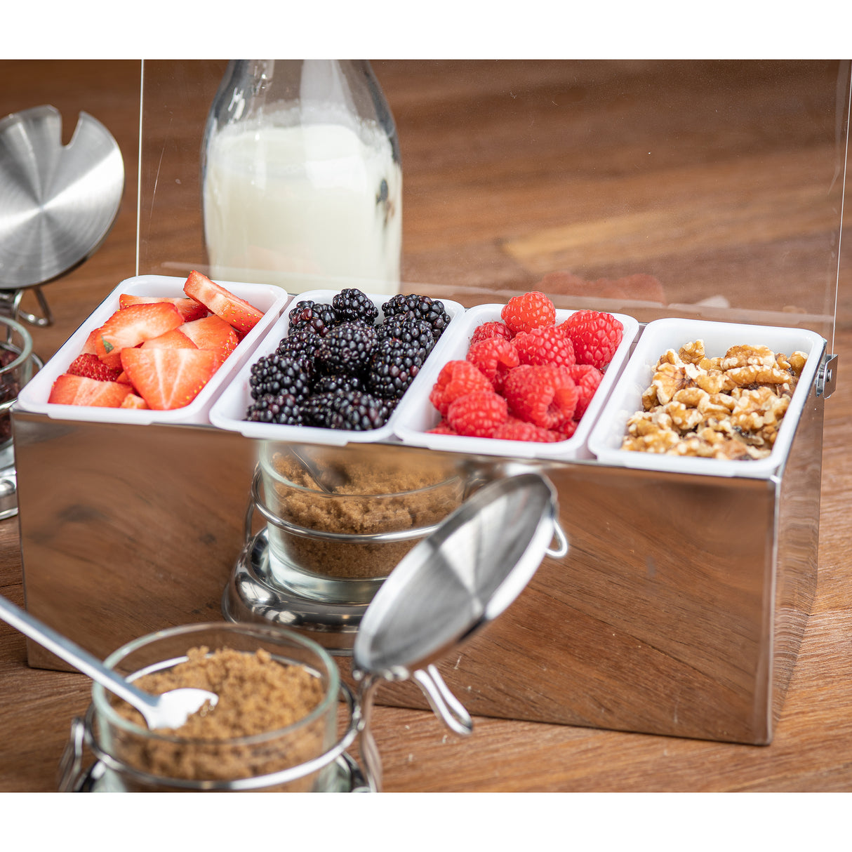 A stainless steel condiment holder with four sections holds sliced strawberries, blackberries, raspberries, and chopped walnuts. Behind it is a bottle of milk, and in front are jars of brown sugar with metal lids and spoons on a wooden surface.