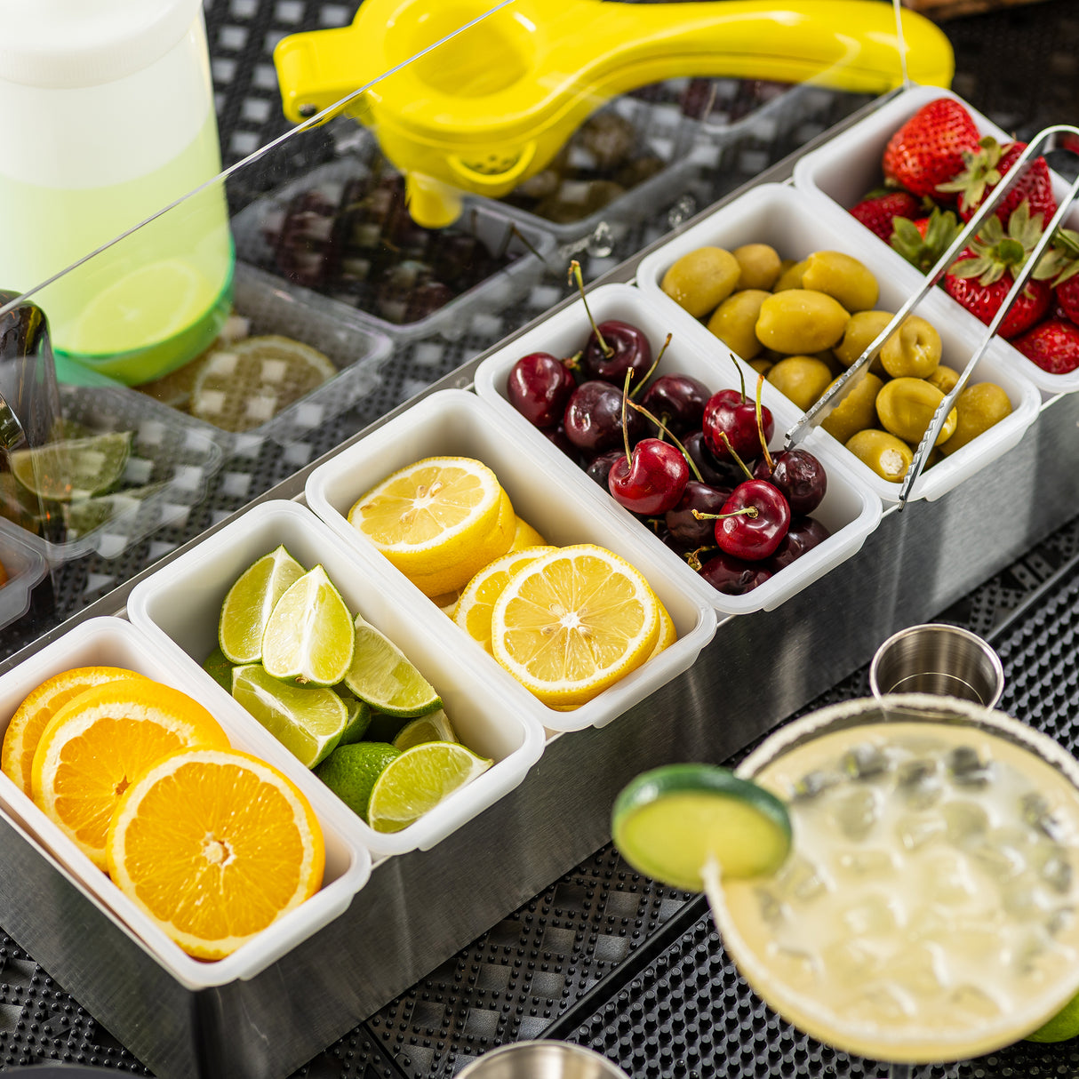 A stainless steel condiment holder with 6 compartments displays orange slices, lime wedges, lemon slices, cherries, green olives, and strawberries. Nearby are a yellow citrus squeezer, a bottle, metal cups, and a creamy drink garnished with cucumber.