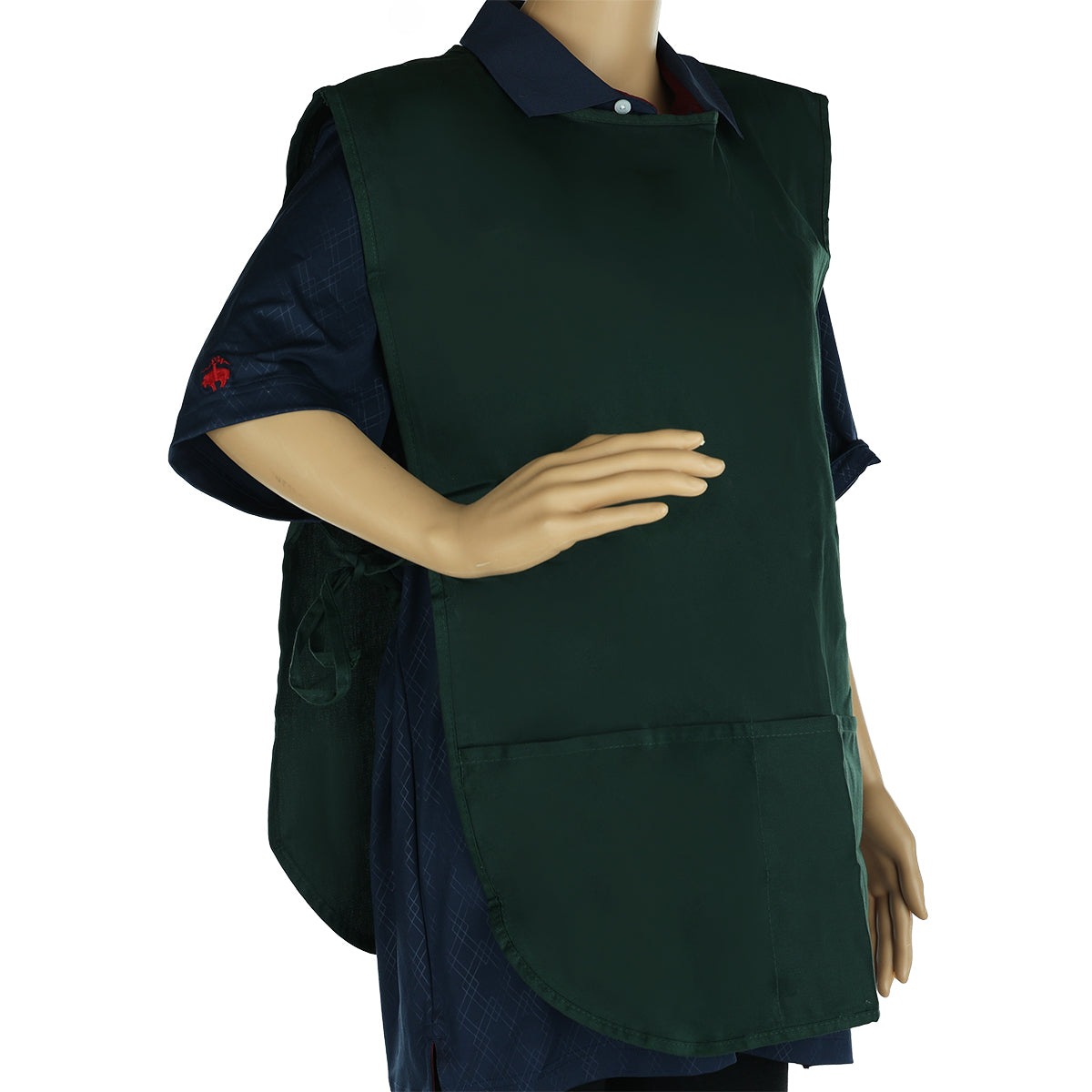 A mannequin dressed in a dark blue collared shirt wears a dark green cobbler apron with two front pockets. Made from a poly-cotton blend, the machine washable apron ties at the back, covering the torso. The left hand rests on its hip; the right arm hangs down.