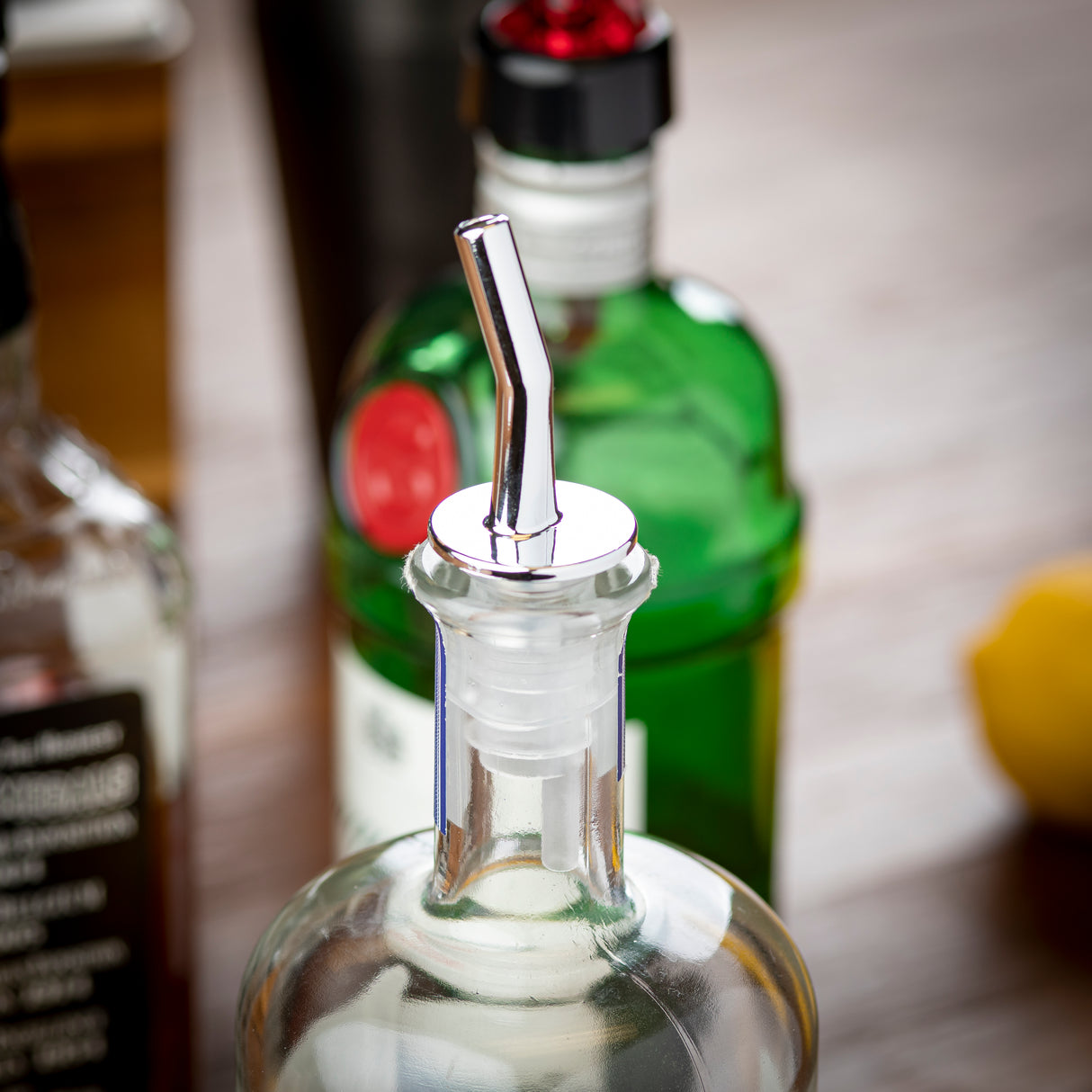 Close-up of a clear glass bottle with a chrome plated spout, likely used as a whiskey pourer. In the blurred background, green and brown glass bottles and a yellow object, possibly a lemon, rest on a wooden surface.