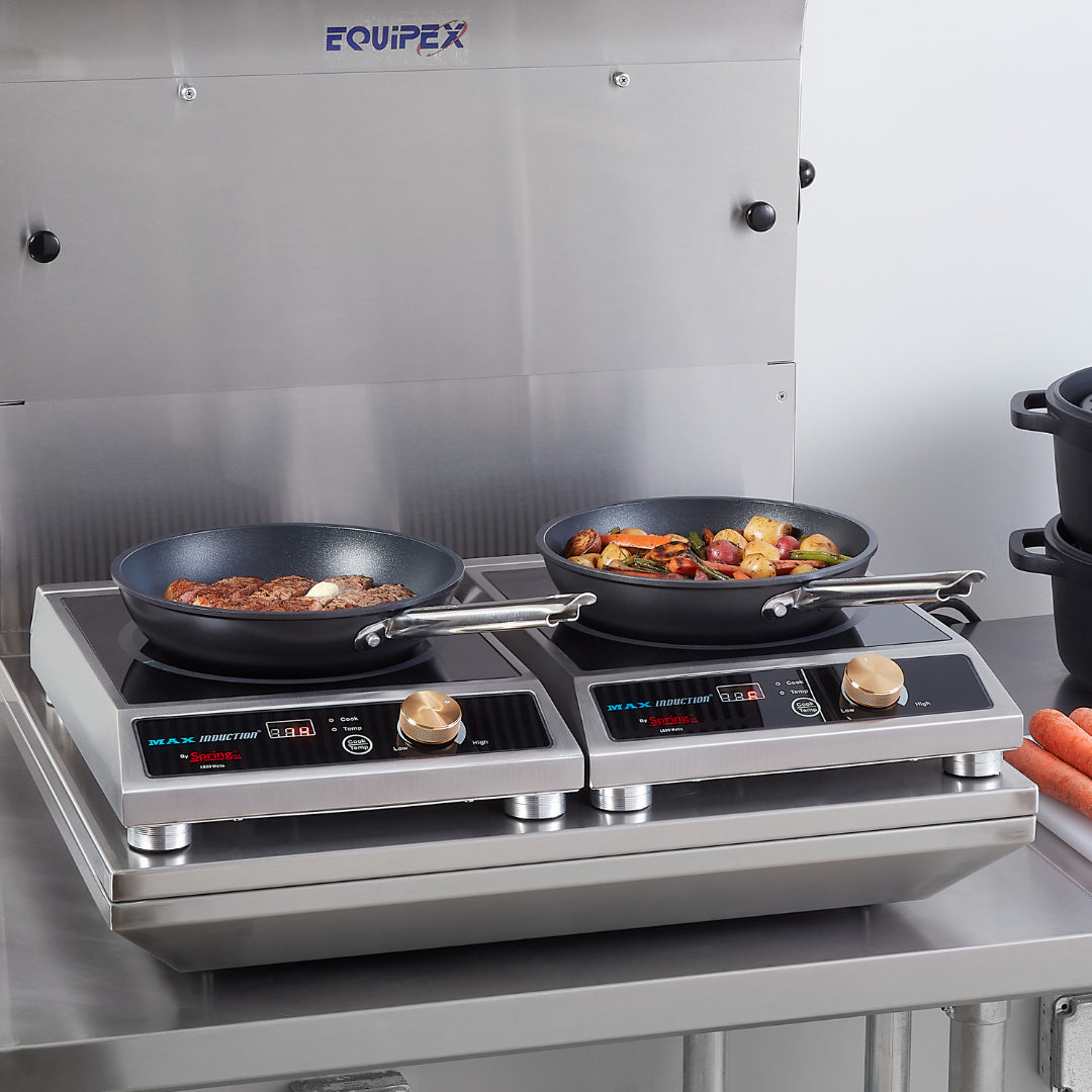 Two frying pans sit on an induction cooktop with digital controls; the left pan holds steak and garlic, while the right contains a colorful vegetable medley. Carrots and stacked black pots rest nearby in a spotless, stainless steel kitchen setting.