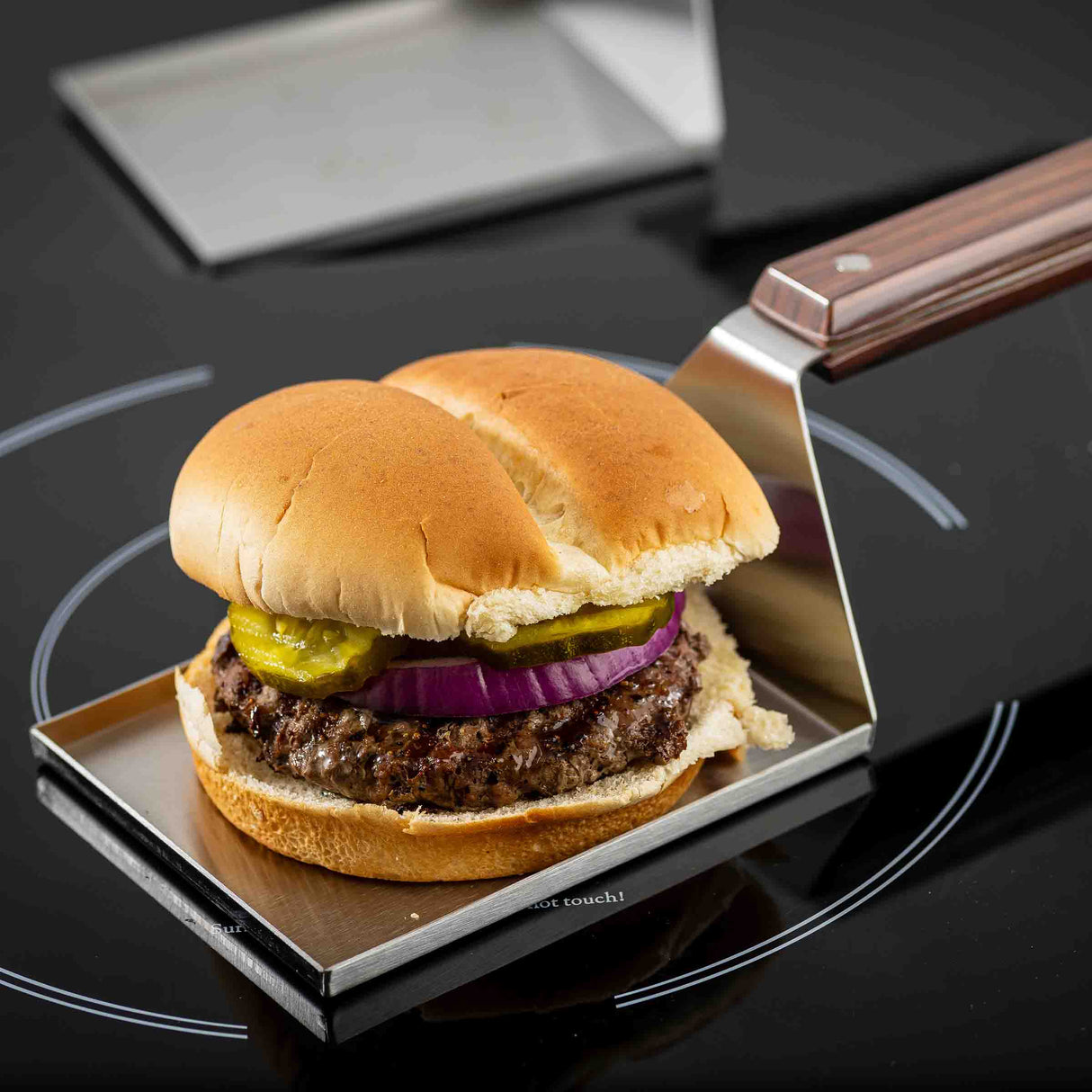 A cheeseburger with pickles and red onion sits on a stainless steel spatula with a wooden handle, resting on a black stovetop. The burger is in a soft, split bun, and the scene is brightly lit, emphasizing the fresh ingredients.