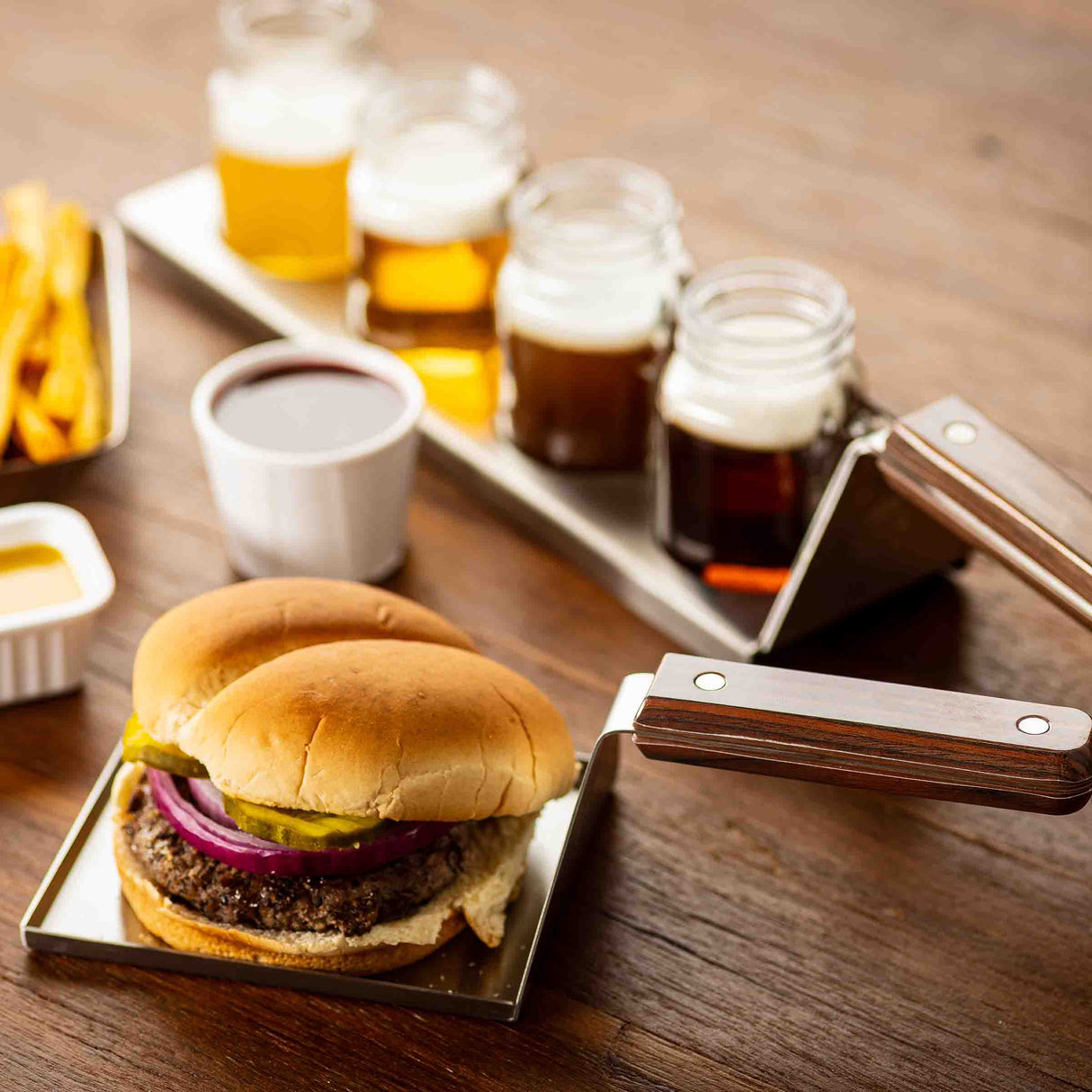 A cheeseburger with red onion, pickles, and a bun sits on a stainless steel spatula beside another bun, with fries, dipping sauces, and four small beer glasses in jars—all set for a casual meal or tasting event. The spatula is dishwasher safe.