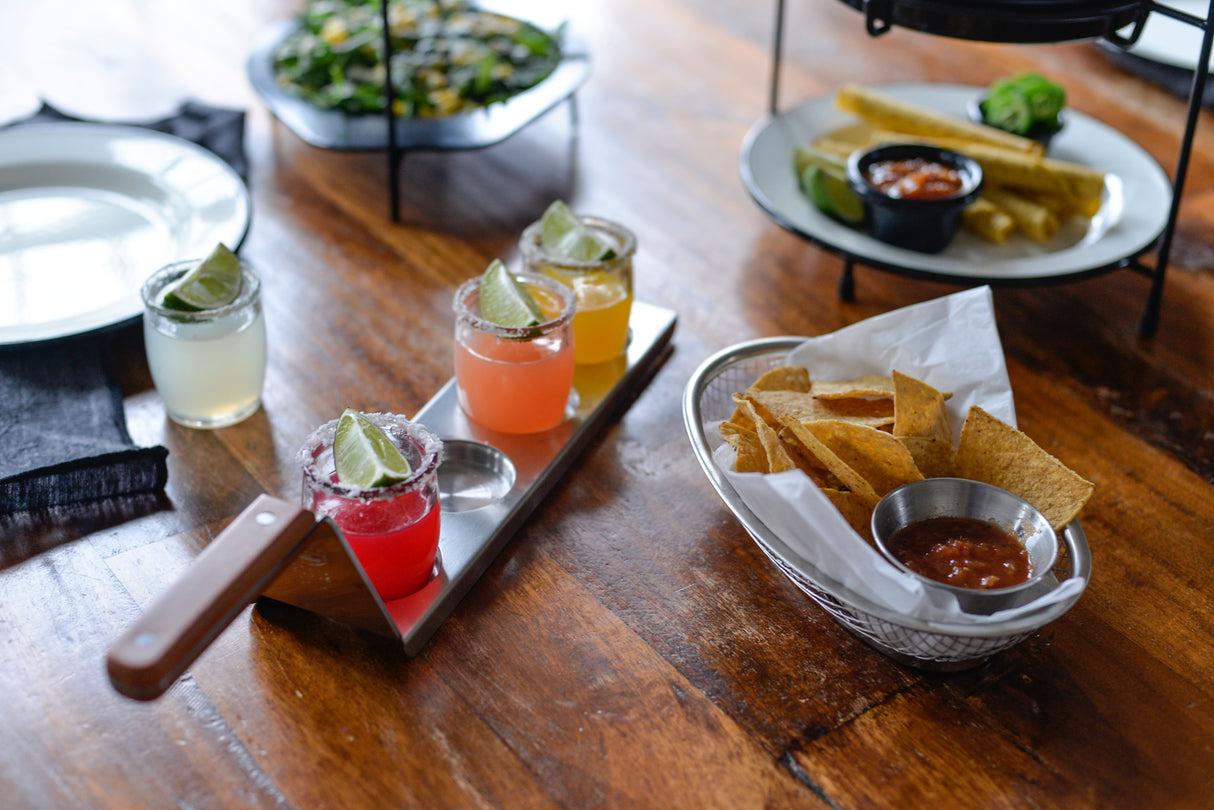 A wooden table with three small cocktails on a stainless steel plate, garnished with lime wedges, a basket of tortilla chips and salsa, taquitos with salsa, and a dish of leafy greens in the background, suggesting a casual dining setting.