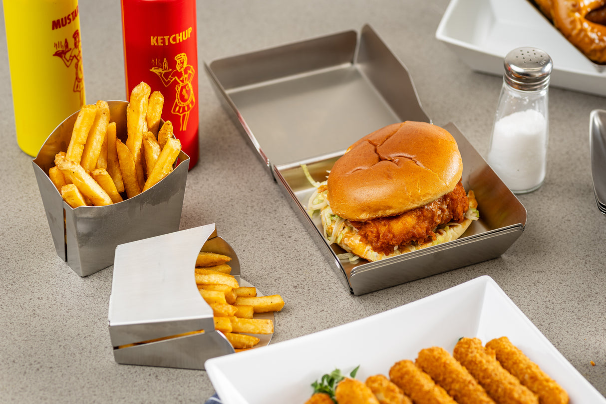 A fried chicken sandwich with lettuce on a bun sits in a stainless steel tray, next to containers of French fries. In the background are bottles of ketchup and mustard, a salt shaker, and mozzarella sticks on a white dish atop a light gray tabletop.