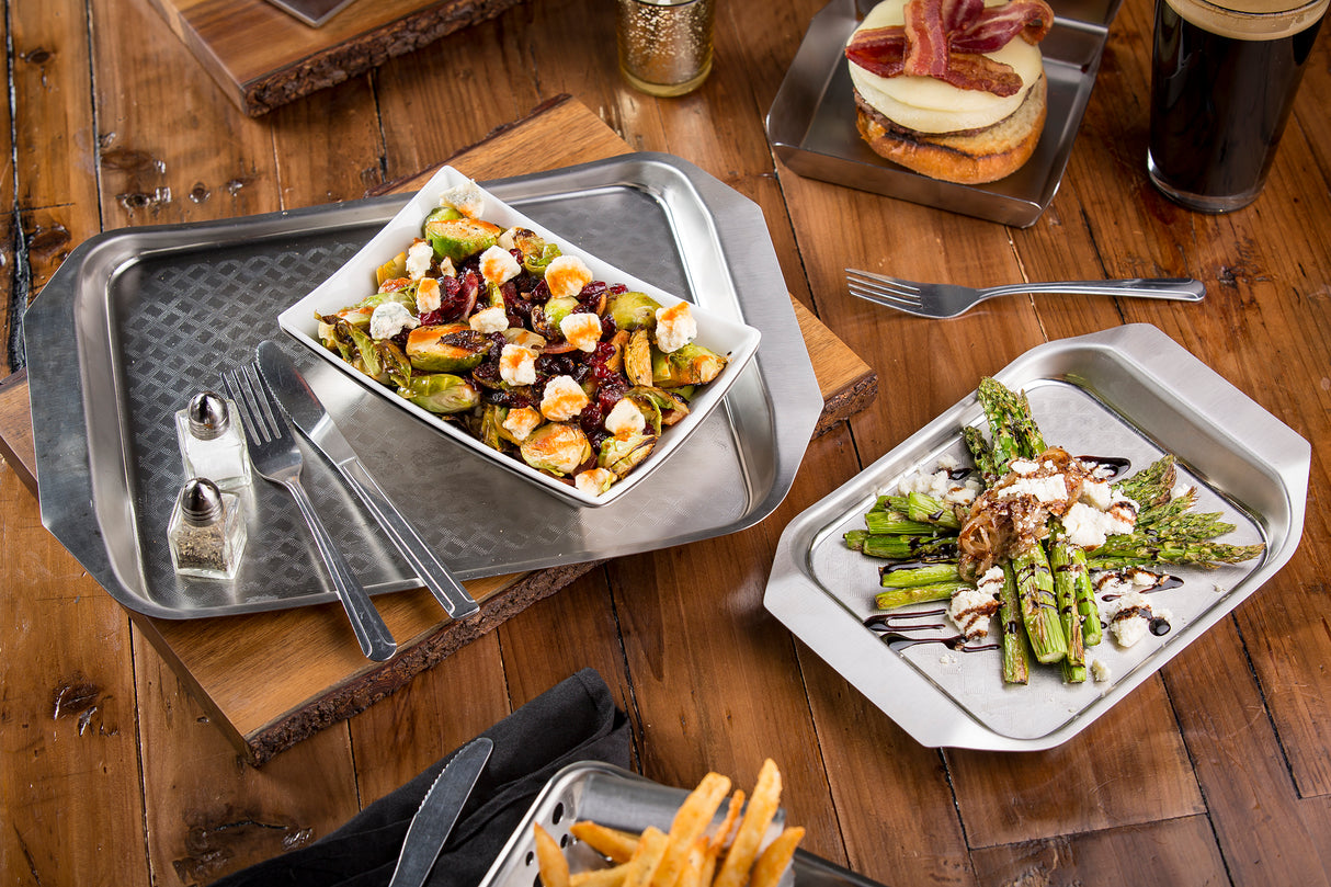 A wooden table features a serving tray with stainless steel metal trays holding roasted brussels sprouts with cheese, grilled asparagus with goat cheese and balsamic drizzle, a Better Burger Collection burger with bacon, fries, and dark beer. Forks and knives are nearby.