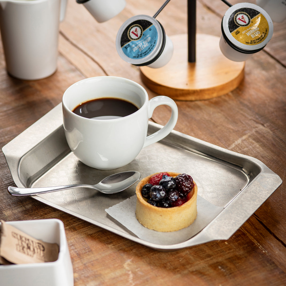 A white mug of black coffee and a small fruit tart topped with berries sit on a stainless steel serving tray with a spoon, on a wooden table. Nearby are coffee pods on a stand and a container with brown sugar packets.
