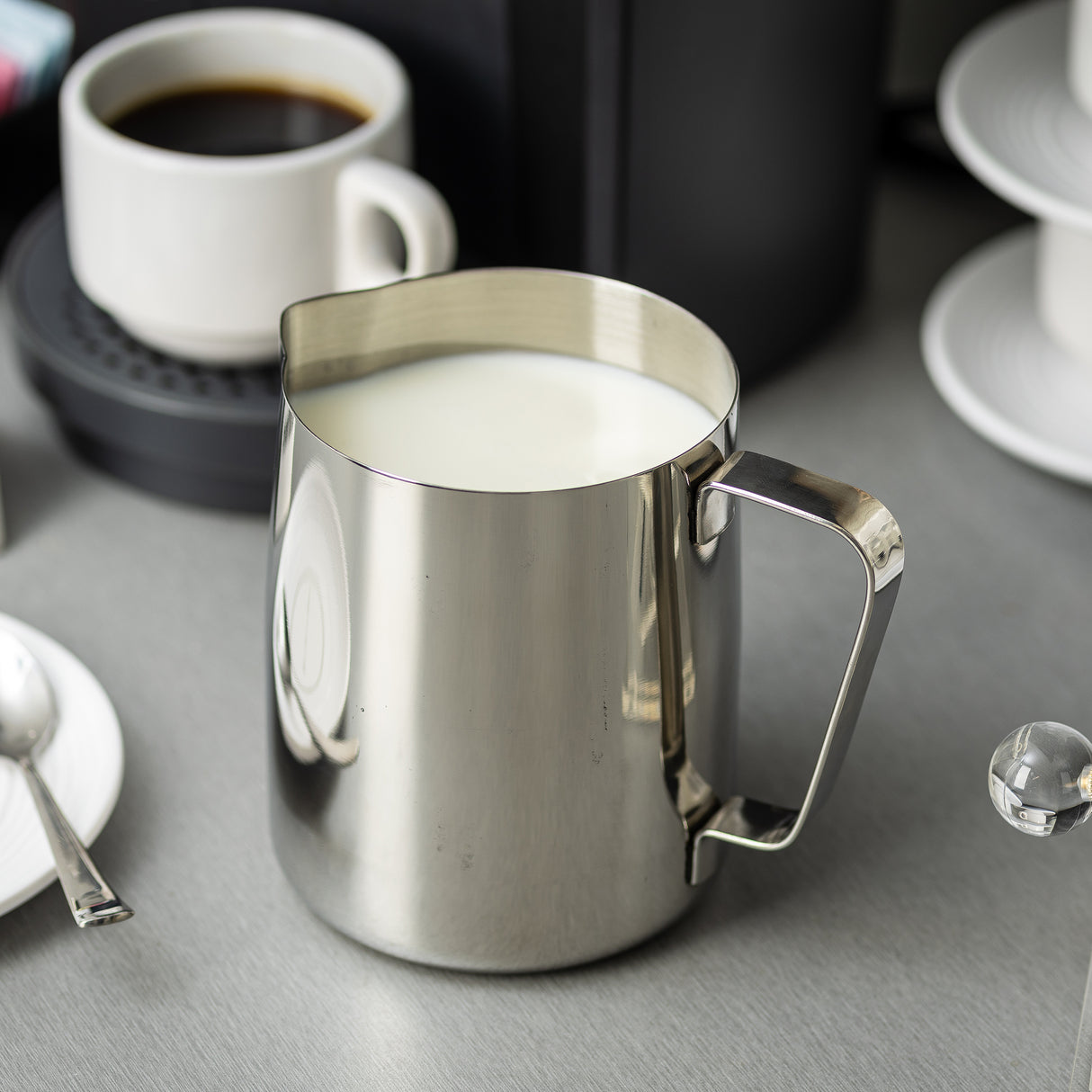 A 36 oz stainless steel frothing cup filled with milk sits on a gray countertop. In the background, there’s a cup of black coffee on a saucer, stacked white cups to the right, and a spoon resting on a white saucer to the left.