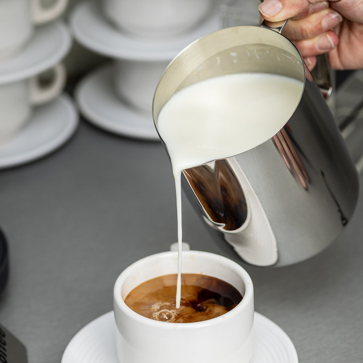 A hand pours creamy milk from a 36 oz stainless steel frothing cup into coffee, creating swirling patterns. The cup sits on a white saucer, with stacked white cups and saucers blurred in the background on a gray countertop.
