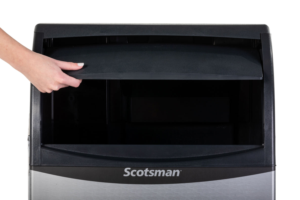 A hand lifts the black lid of a Scotsman Ice Maker, revealing the dark, empty interior. The metallic front displays the Scotsman logo, showcasing this undercounter ice maker against a plain white background.