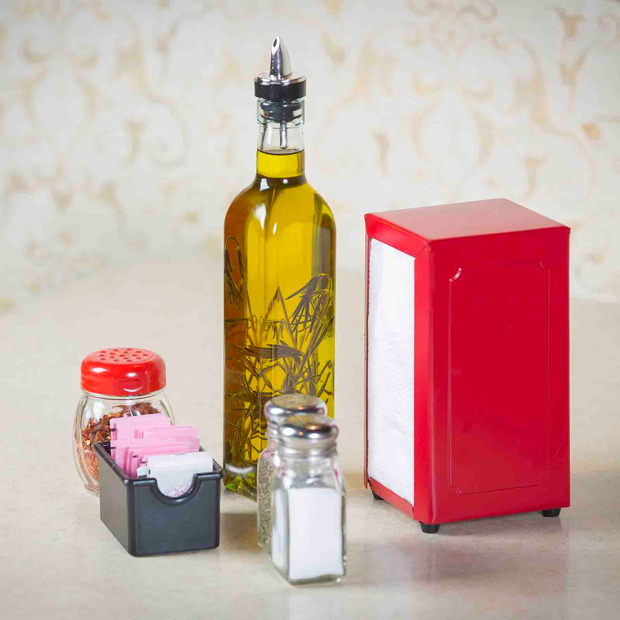 A bottle of olive oil with a spout, a TableCraft stainless steel napkin dispenser, a small jar of crushed red pepper, a black container holding pink and white sweetener packets, and salt and pepper shakers are arranged on a light countertop.