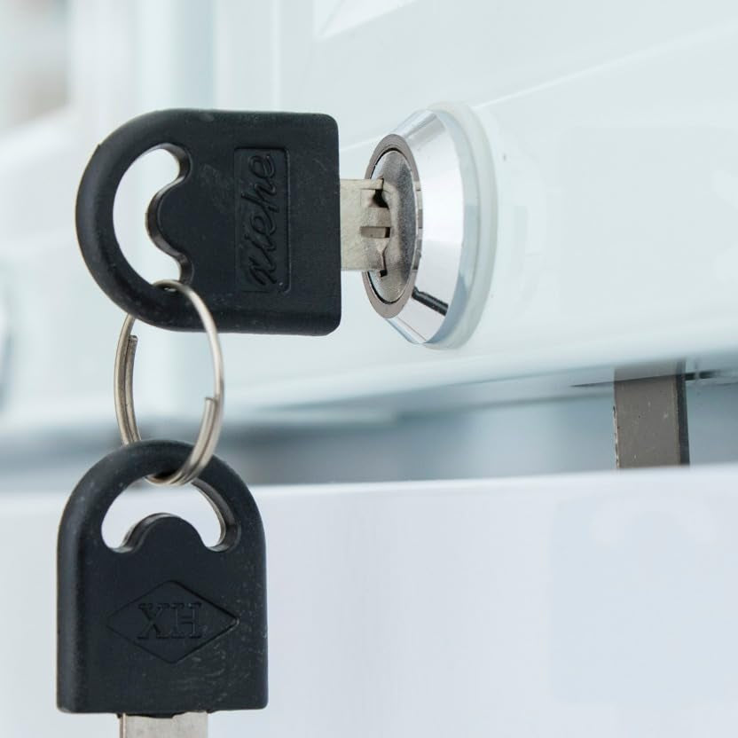 Close-up of a metal key inserted into a cylindrical lock on a stainless steel refrigerator, with another identical key hanging from the ring. Both keys have black plastic heads and the lock is mounted on a white glossy surface.