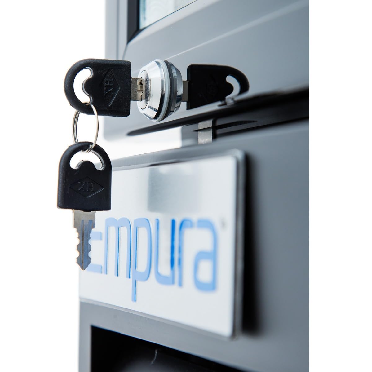 Close-up of a lock with black keys inserted, set on a metallic gray surface. Below, a white and blue sign partially shows “Empura,” hinting at Empura Refrigeration—perfect for securing your Glass Door Cooler or Refrigerated Merchandiser.