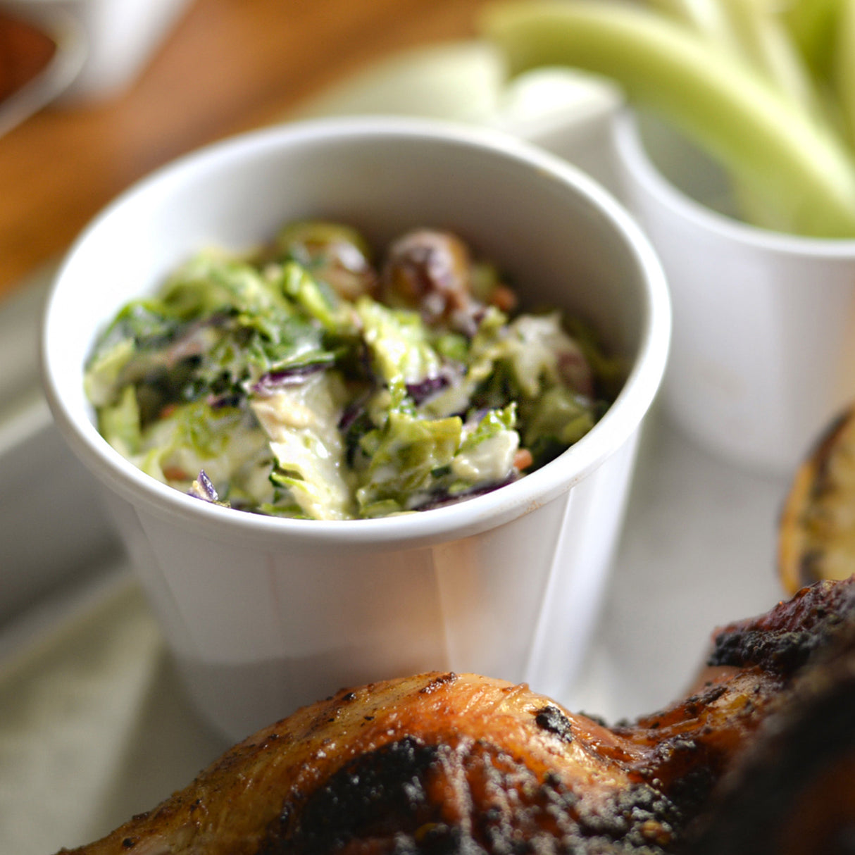 A white melamine soufflé cup filled with creamy coleslaw, featuring chopped cabbage and carrots, sits on a tray next to grilled meat and sliced celery in another cup. The coleslaw appears fresh and colorful, with visible herbs mixed in.